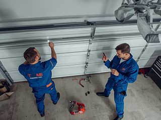 Two professional technicians in blue uniforms servicing a residential garage door, surrounded by tools, highlighting expert maintenance and repair services.
