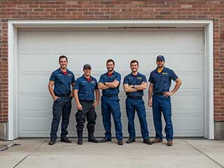 A crew of uniformed garage door technicians standing confidently in front of a brick residence with a white garage door, symbolizing expert local service and reliability.