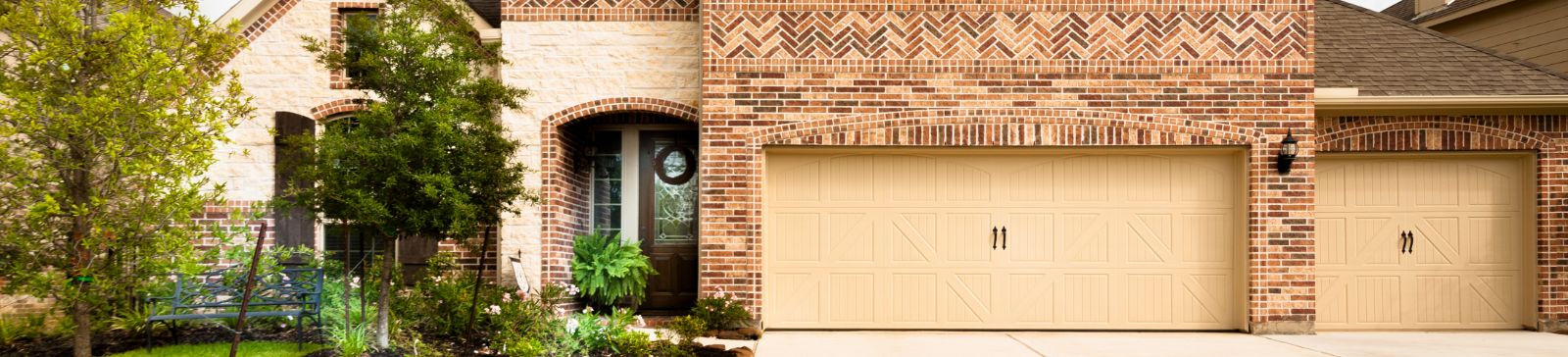 Front view of a brick house featuring three beige garage doors, custom brickwork, and well-maintained landscaping, ideal for showcasing quality garage door repair and installation services.
